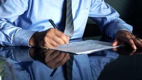 Businessman Writing Notes In Documents Sitting By Glass Table In Office