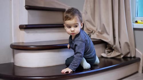 Cute little boy goes down by the stairs indoors. Toddler moves backward carefully watching his way.