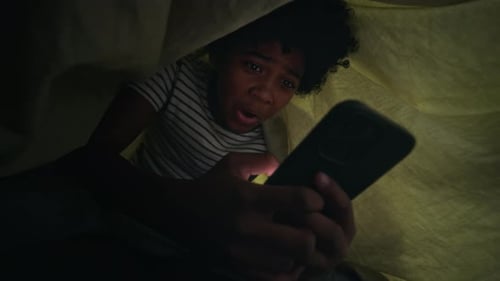 Garçon joyeux se cachant sous une couette et regardant du contenu divertissant la nuit