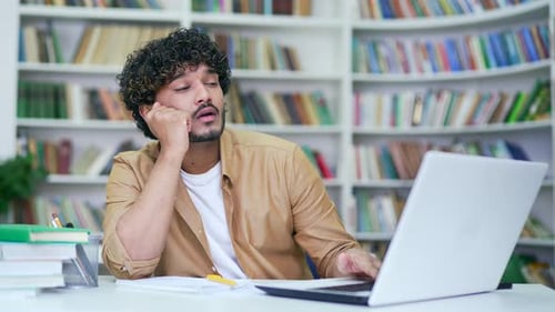 Tired student studying using laptop in campus library space. Exhausted male learning on examination