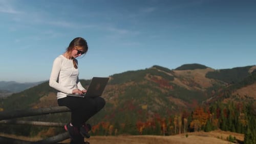 Woman Using Laptop Sitting on Wooden Fence in Mountains