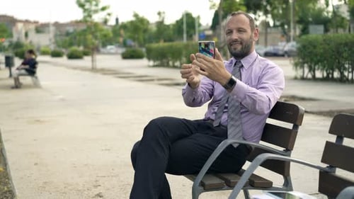 Young, Happy Businessman Taking Photo with Cellphone on Bench in City 30s