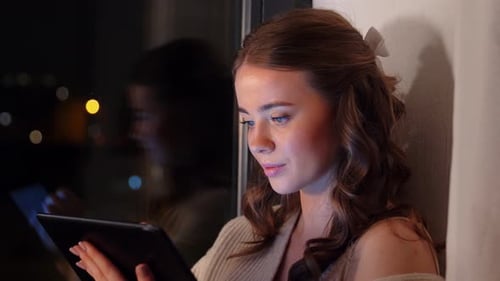 Young woman browsing tablet at window in cozy sweater on winter night