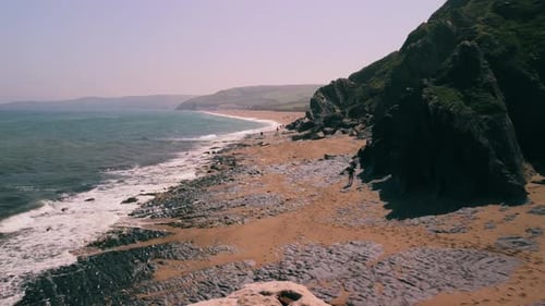 Drone shot moving along a beach in the summer as people walk on the sand. Cliffs to the right and th