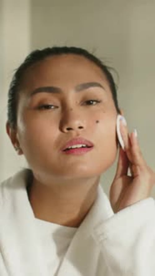 Vertical video: Starting mid adult Asian woman removing makeup in robe at home, showing cotton pads