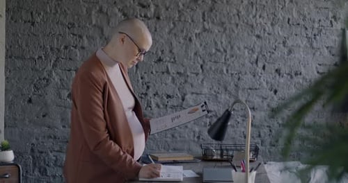 Side View of Pregnant Woman Entrepreneur Writing in Notebook and Reading Business Document in Office