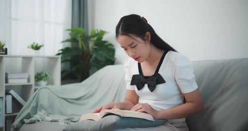 Junge asiatische Frau sitzt entspannt auf der Couch und liest ein Buch im Wohnzimmer.