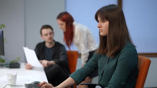 Side View Concentrated Young Woman Working in Office with Blurred Colleagues Talking at Background