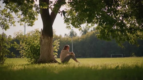 Thoughtful Woman Sitting on Grassy Ground in Sunlit Park
