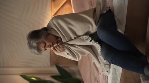 Une femme âgée est assise dans son lit enveloppée dans une couverture et a l'air malade, elle a probablement attrapé un rhume ou