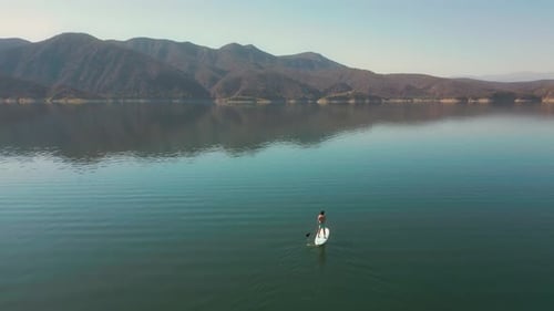 Aerial drone flying over a lake, man doing stand up paddle, with mountains at sunrise. SUP.