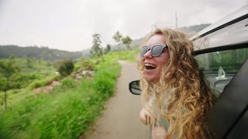 Cheerful Woman with Curly Hair Enjoys Mountain Road Trip in Car Smiling Female Traveler Leans Out