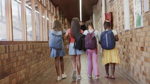 Rear view of happy diverse schoolgirls with school bags walking in corridor at elementary school