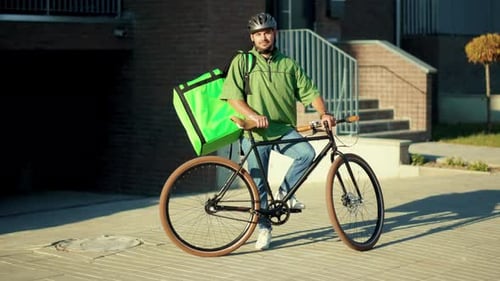 Au soleil, portrait d'un livreur attrayant debout avec un sac à dos de vélo regardant la caméra