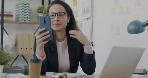 Middle Eastern Businesswoman Talking on Video Call Using Smartphone and Earphones in Office