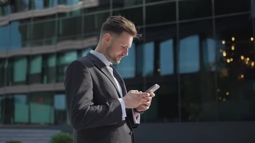Young Businessman Holding Phone Reading Email Stands on Street Smiling Writes Message Communicates