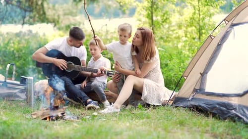 camping by lake in woods. happy family dad mom and little kids sitting by fire and tent in nature.