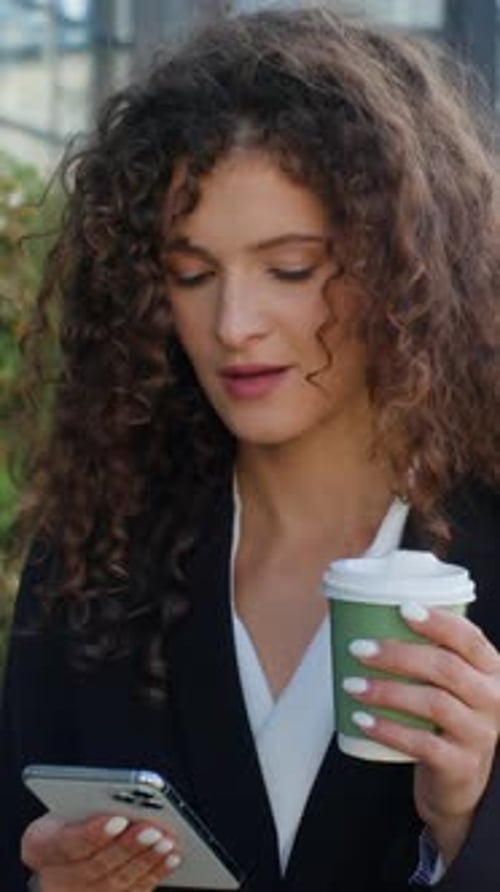 Mujer de negocios caucásica adicta a los artilugios que busca un teléfono móvil con un café caminando por la ciudad