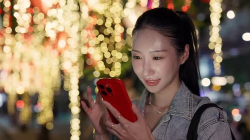 Portrait of Happy Asian Woman Reads Messages on Smartphone Against Bokeh Lights
