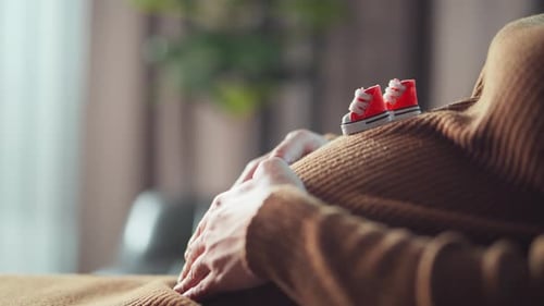 Pregnent woman hands caress her belly while a pair of small kid shoe is placed nearby