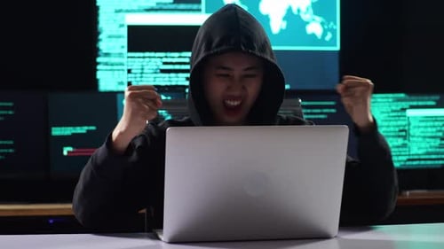 Hacker asiática em um capuz comemorando enquanto usa o laptop com vários monitores em um escritório escuro