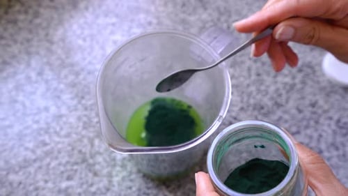 Adding And Stirring A Small Spoonful Of Spirulina Powder To Green Juice In A Pitcher. overhead