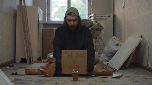 Homeless Poor Man Sitting in a Room of an Abandoned Building with a Money Jar He is Holding a Piece