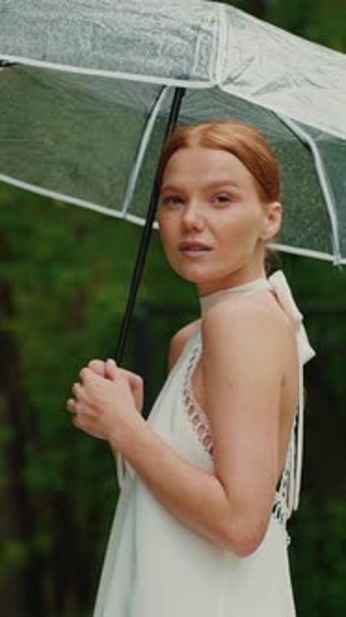 Young Woman Smiles While Holding a Clear Umbrella in the Rain at a Lush Green Park