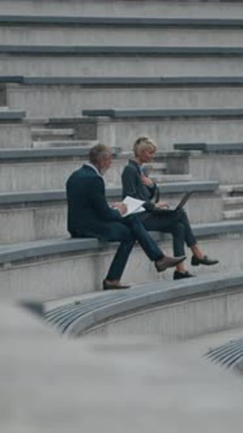 Businesspeople Sitting on City Stairs Using Laptop and Discussing Documents