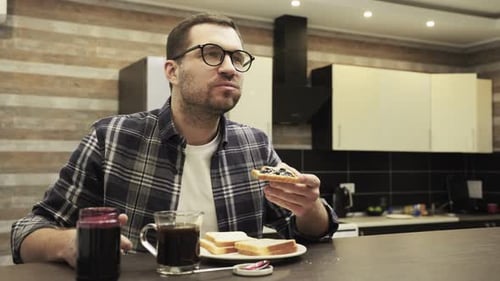 Young Bearded Guy in Glasses Having Breakfast and Biting Toast with Jam at Home