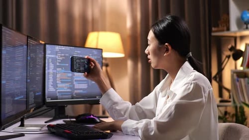 Woman Programmer Looking At Desktop Computer Screen And Using Smartphone While Creating Software