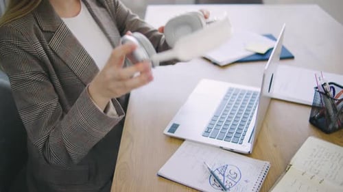 Woman Using Headphones While Working at Her Desk with a Laptop and Notebooks in a Modern Office