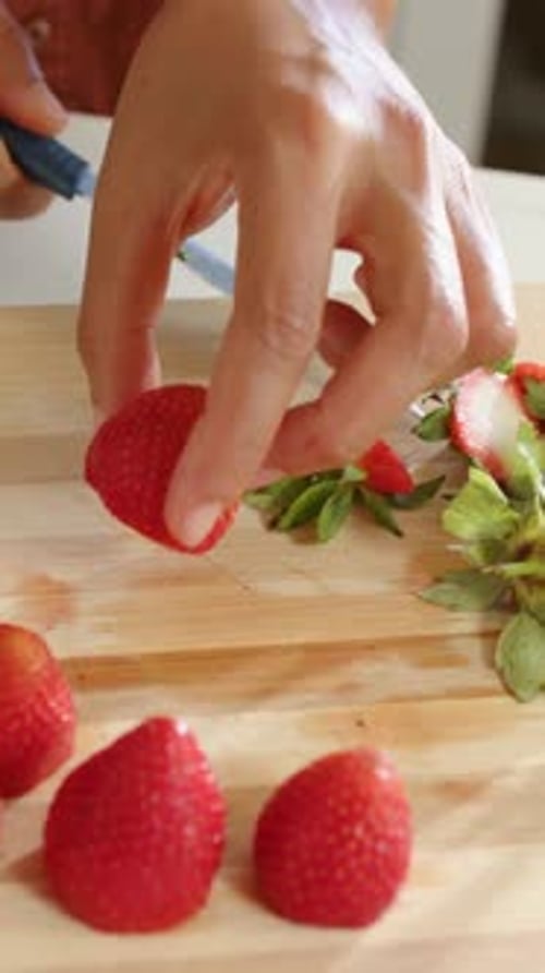 Vertical video: Sliding blue knife under stem, woman in orange paring berries at counter to stand