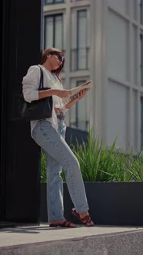 Young Woman Reading a Newspaper While Leaning Against a Building Pillar