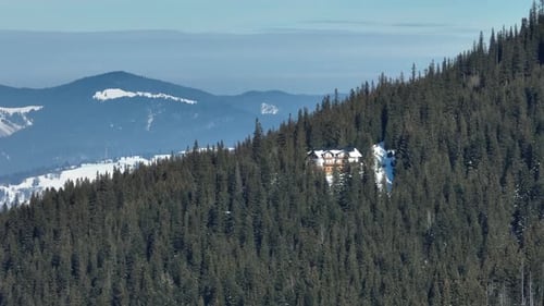 A snowy lodge nestled in a dense evergreen forest on a mountain slope with distant peaks
