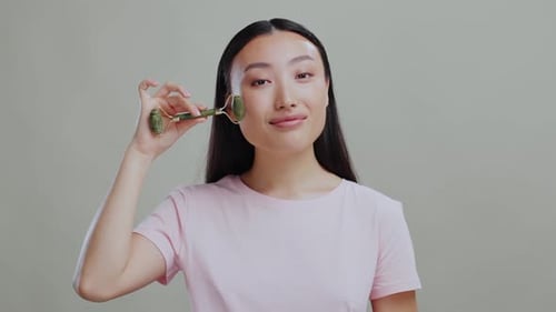 Young Attractive Japanese Woman in Grey Studio Background Doing Gentle Face Massage Using Jade