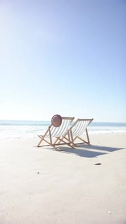 Vertical video: Two beach chairs with hat on sandy shore, relaxing by ocean