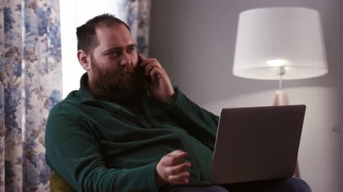 A Man in a Green Sweater Sits in a Chair and Talks on the Phone A Bearded Male Freelancer Sits in a