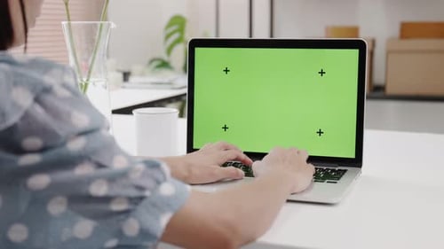 Over the Shoulder : Young Woman Sitting at Her Desk Using Laptop with Mock-up Green Screen.