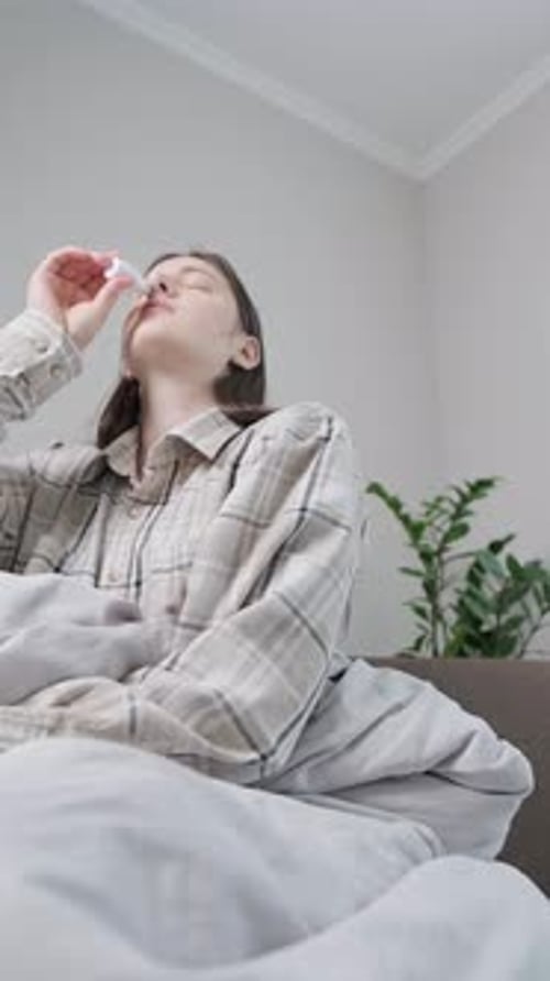 Jeune femme recouverte d'une couverture allongée sur le canapé à la maison, utilisant un spray nasal, traitement à domicile pour