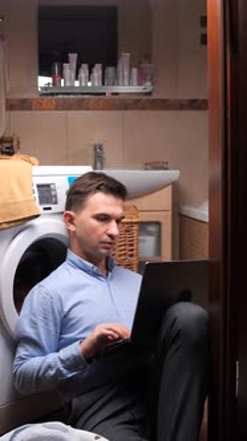 Vertical Video Man Sitting on the Floor in the Bathroom Using a Laptop While Waiting for the Washing