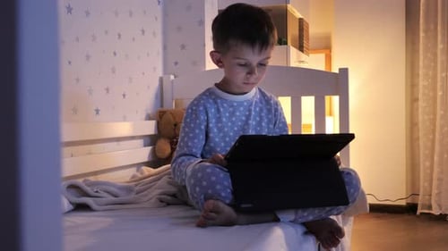 Little boy in pajamas sitting with tablet computer on bed and watching video