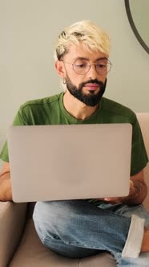 Young blond man with a beard works on a laptop while sitting in a cozy living room