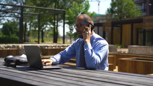Businessman With Laptop Talking On Cellphone Sitting In City Park