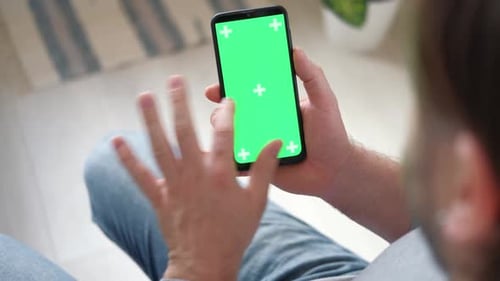 Young Man Sitting at Home Holding Smartphone Green Mockup Screen in Hand Male Person Using Chroma