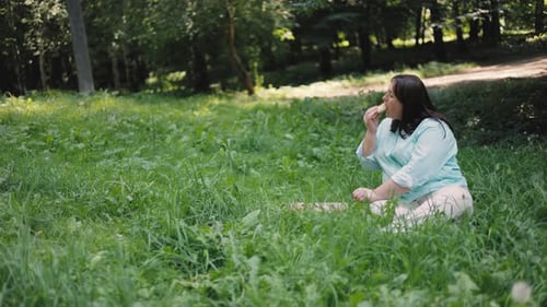 A brown-haired girl in a striped shirt sits on the grass in the park and eats pizza