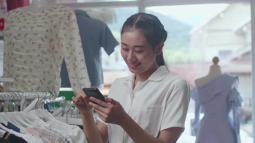 Asian Woman Using Smartphone While Choosing Various Clothing Items On Hangers At The Boutique