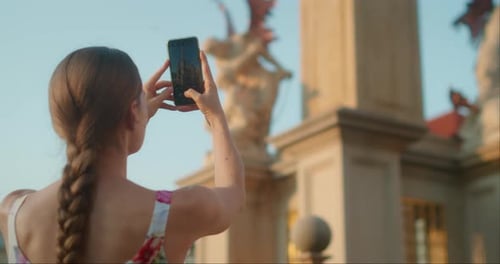 Woman photographs a historic statue near a grand architectural gate in Prague during summer travel