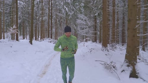 Athletic man in green outfit running in woods on snowy trail during cold winter day - follow shot, s