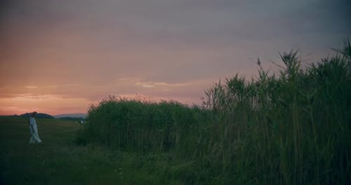 Woman Walking Towards Grass In Meadow At Dusk